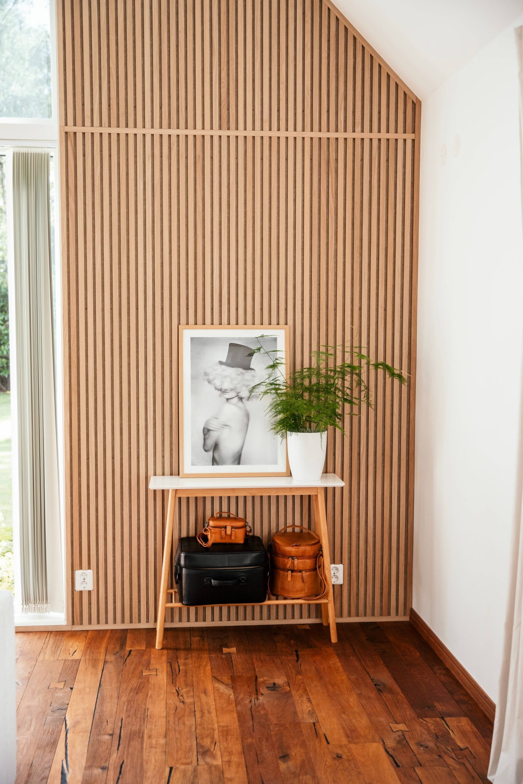 Une petite table en bois contenant des sacs, une photo encadrée et une plante en pot est placée contre un panneau acoustique nordique dans une pièce lumineuse avec des planchers en bois.